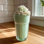 A tall glass filled with a bright green Copycat Shamrock Shake topped high with mint whipped cream.