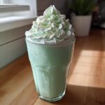 A tall glass of green Copycat Shamrock Shake topped with mint whipped cream and gold sprinkles.