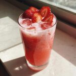 Tall glass filled with a bright pink, frothy Copycat Starbucks Pink Drink, topped with ice and sliced strawberries.