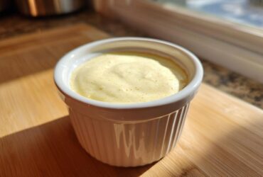 A small white ramekin filled with creamy yellow Copycat Taco Bell Chicken Quesadilla Sauce, sitting on a wooden board.