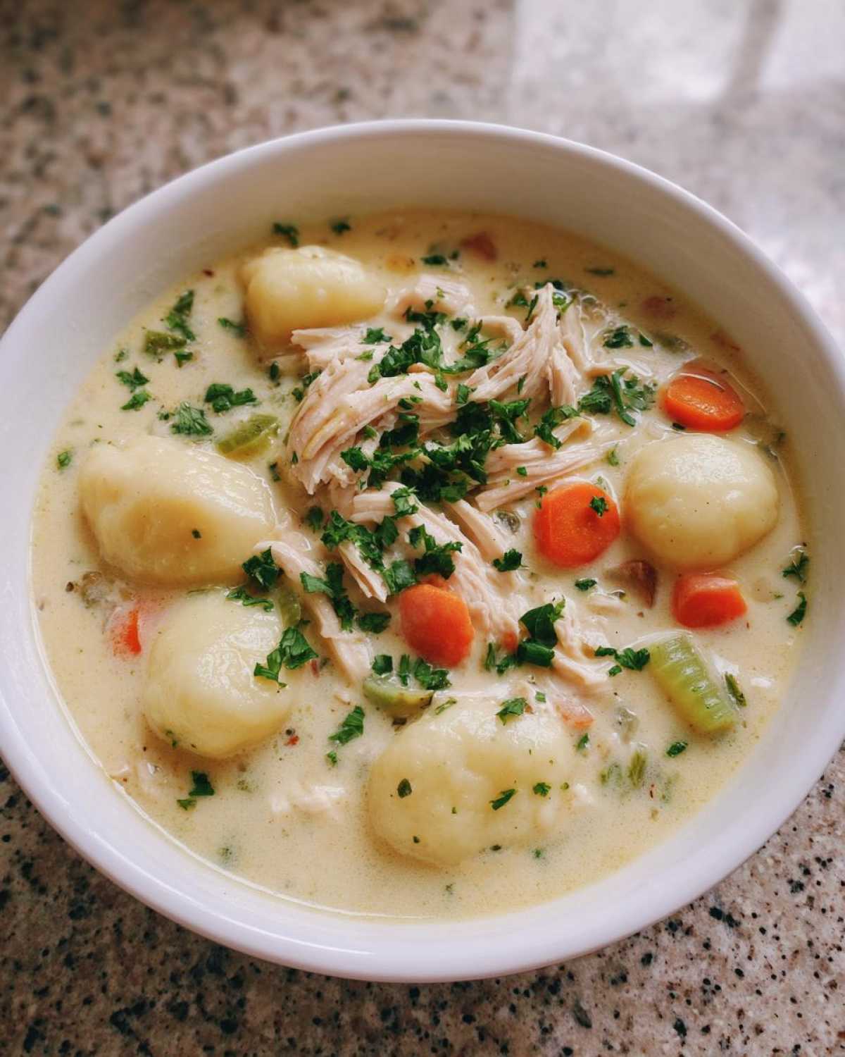 A close-up of a white bowl filled with rich Creamy Chicken Gnocchi Soup, topped with shredded chicken and fresh parsley.