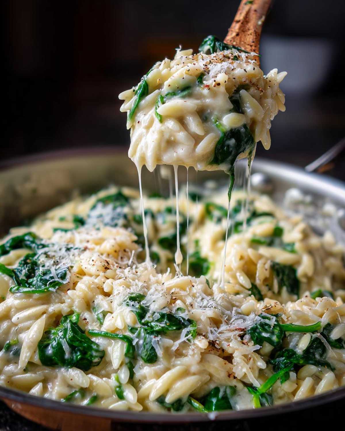 A wooden spoon lifts a scoop of creamy garlic parmesan orzo with spinach, showing cheese pulls.