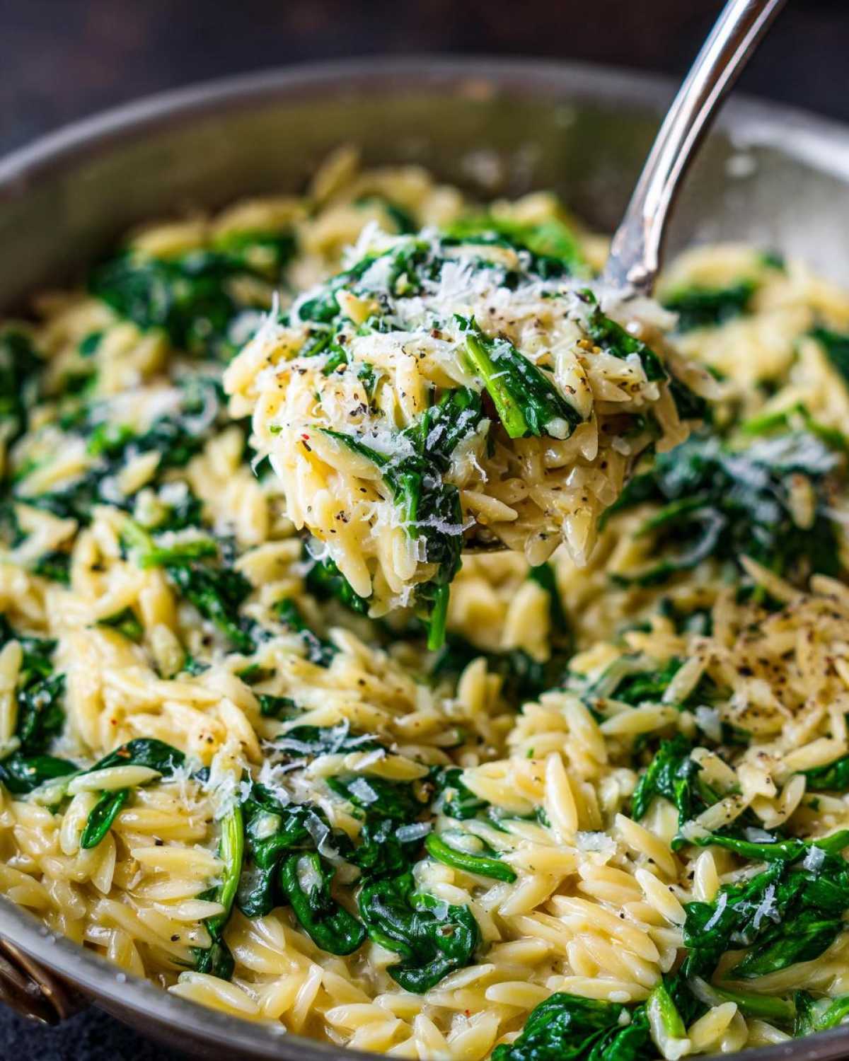 A spoonful of creamy garlic parmesan orzo with spinach being lifted from the pan, topped with grated cheese.