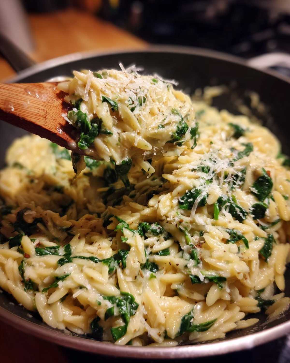 A wooden spoon lifts a serving of Creamy Garlic Parmesan Orzo with Spinach topped with grated cheese.