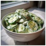A white bowl filled with slices of Creamy German Cucumber Salad generously coated in a dill-flecked white dressing.