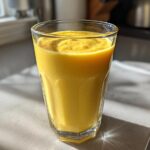 A tall glass filled with a vibrant, creamy yellow Pineapple Coconut Smoothie sitting on a countertop in bright sunlight.