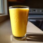 A tall glass filled with a thick, bright yellow Pineapple Coconut Smoothie sitting on a kitchen counter in sunlight.