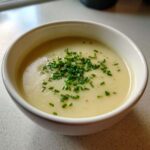 A close-up of a white bowl filled with smooth, creamy potato leek soup, topped generously with fresh chopped chives.
