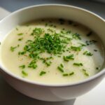 Close-up of a white bowl filled with smooth Creamy Potato Leek Soup, topped with fresh chopped chives and pepper.