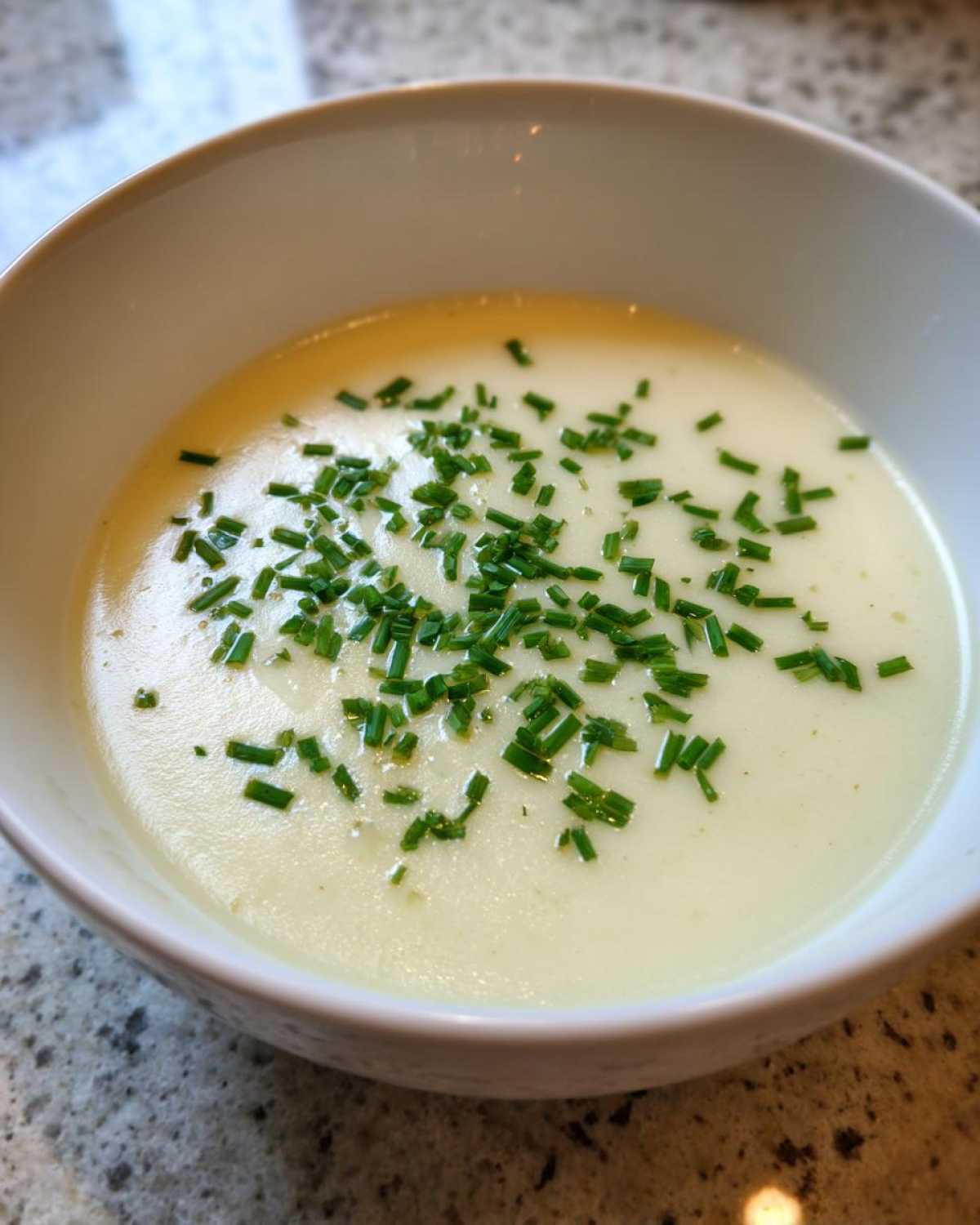 A close-up of a white bowl filled with smooth, creamy potato leek soup, garnished generously with chopped green chives.