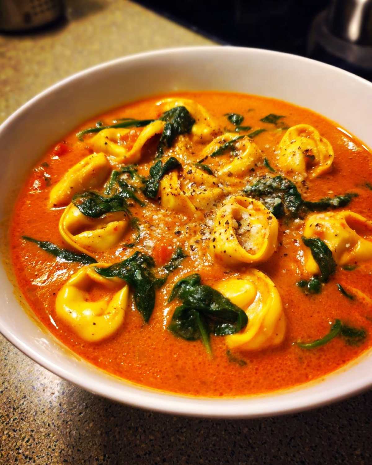 A close-up of a white bowl filled with rich Creamy Tomato Tortellini Soup, featuring cheese tortellini and wilted spinach, topped with Parmesan.