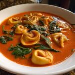 A close-up of a white bowl filled with rich, orange Creamy Tomato Tortellini Soup, featuring tortellini and wilted spinach.