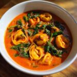A close-up of a white bowl filled with rich Creamy Tomato Tortellini Soup featuring cheese tortellini and wilted spinach.