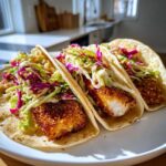 Three Crispy Baked Fish Tacos loaded with crunchy lime slaw on a white plate.
