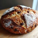 A rustic, round loaf of Easy Irish Soda Bread with buttermilk and raisins, featuring a golden-brown, cracked crust dusted with flour.