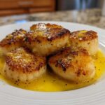Close-up of five golden-brown seared Garlic Butter Scallops with Lemon resting in bright yellow butter sauce on a white plate.