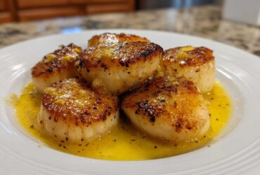 Close-up of five golden-brown seared Garlic Butter Scallops with Lemon resting in bright yellow butter sauce on a white plate.