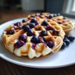 Close-up of golden Gluten-Free Blueberry Waffles topped with fresh blueberries and syrup on a white plate.