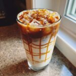 A tall glass of Iced Caramel Macchiato showing layers of milk, ice, and rich caramel sauce drizzled inside the glass.