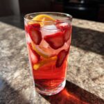 Tall glass filled with vibrant red Iced Strawberry Lemon Tea, ice, sliced strawberries, and lemon wedges.