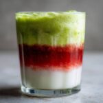 Close-up of a vibrant Iced Strawberry Matcha Latte showing distinct layers of white milk, red strawberry puree, and green matcha foam.