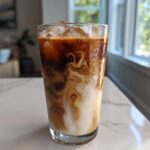 A tall glass of Iced Vanilla Chai Latte showing rich swirls of dark chai concentrate mixing with white milk over ice.