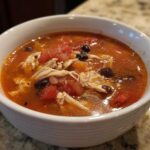 Close-up of a white bowl filled with rich, red broth Instant Pot Chicken Tortilla Soup, featuring shredded chicken, black beans, and diced tomatoes.