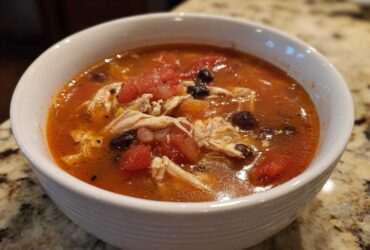 Close-up of a white bowl filled with rich, red broth Instant Pot Chicken Tortilla Soup, featuring shredded chicken, black beans, and diced tomatoes.