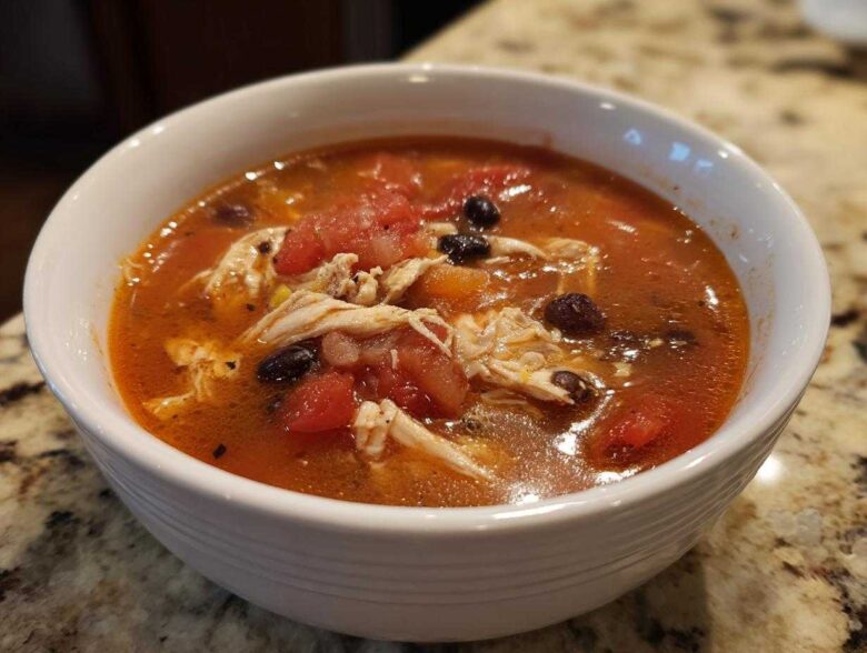 Close-up of a white bowl filled with rich, red broth Instant Pot Chicken Tortilla Soup, featuring shredded chicken, black beans, and diced tomatoes.