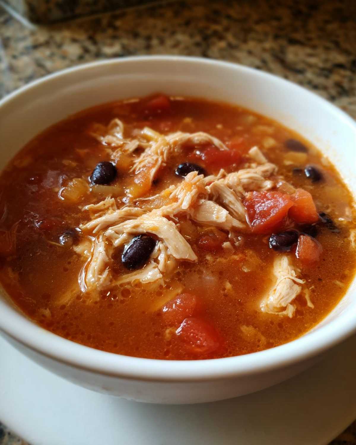 A close-up view of a white bowl filled with rich, red broth Instant Pot Chicken Tortilla Soup, featuring shredded chicken and black beans.