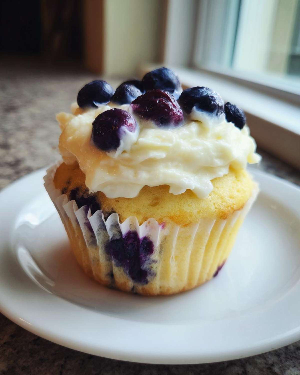 A single Lemon Blueberry Cupcake with thick cream cheese frosting topped with fresh blueberries, sitting on a white plate.