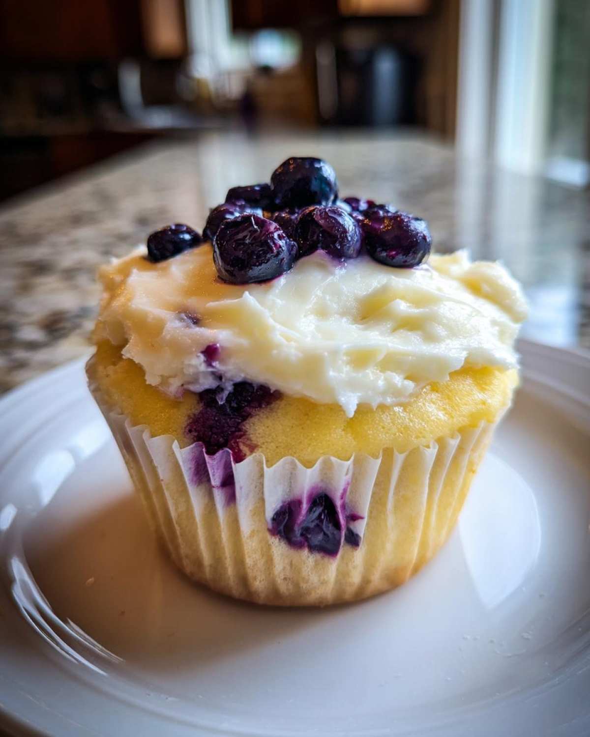 A single Lemon Blueberry Cupcake with Cream Cheese Frosting, topped with fresh blueberries, sitting on a white plate.