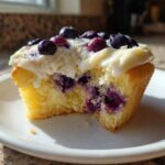 Close-up of a Lemon Blueberry Cupcake with cream cheese frosting, showing a bite taken out revealing moist cake and blueberries.