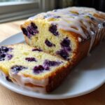 A moist Lemon Blueberry Yogurt Loaf drizzled with white glaze, with one slice cut and resting beside the main loaf.