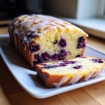 A moist Lemon Blueberry Yogurt Loaf, drizzled with white glaze, with one slice cut and resting beside the main loaf.