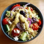 Close-up of a Mediterranean Quinoa Bowl topped with feta, olives, tomatoes, red onion, and lemon tahini sauce.