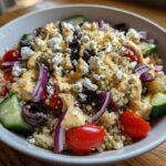 Close-up of a vibrant Mediterranean Quinoa Bowl topped with feta, olives, tomatoes, cucumber, and lemon tahini sauce.