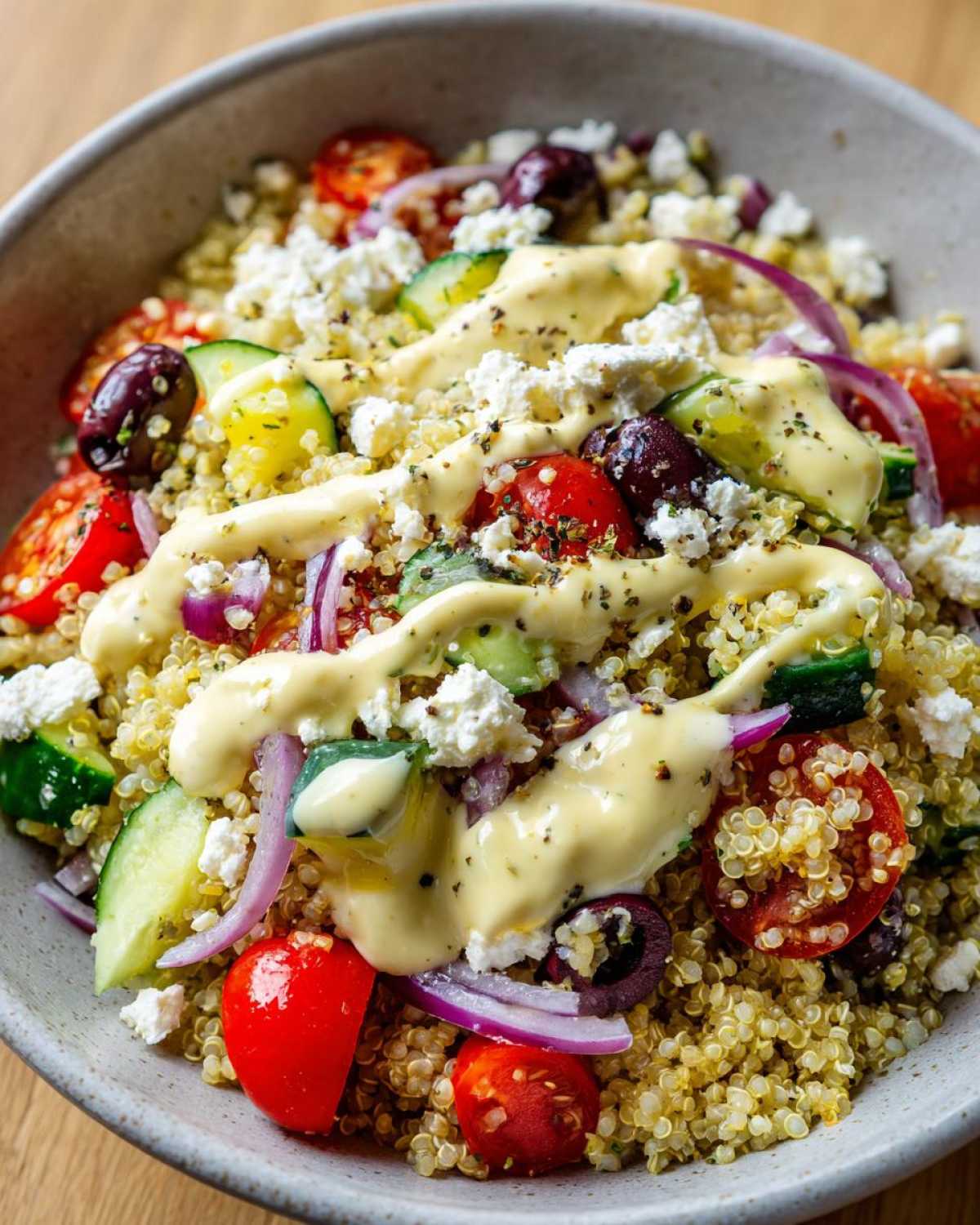 Close-up of a Mediterranean Quinoa Bowl topped with feta, cucumbers, tomatoes, olives, and lemon tahini sauce.