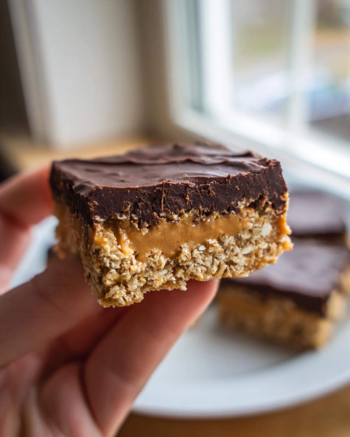 A hand holding a layered No-Bake Chocolate Peanut Butter Oat Bars showing oat base, peanut butter filling, and chocolate topping.