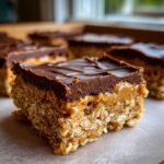 A close-up of a thick No-Bake Chocolate Peanut Butter Oat Bars slice showing the oat base, peanut butter layer, and chocolate topping.