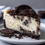 A rich slice of No-Bake Oreo Cheesecake topped with cookie crumbs and a whole Oreo cookie beside it.