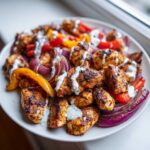 Close-up of seasoned Sheet Pan Chicken Shawarma pieces topped with garlic yogurt sauce and roasted vegetables.