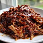 A close-up of shredded Slow Cooker Honey Garlic Pulled Pork, glistening with rich, dark sauce on a white plate.