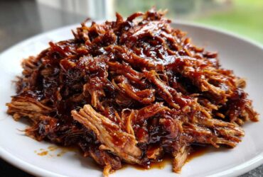 A close-up of shredded Slow Cooker Honey Garlic Pulled Pork, glistening with rich, dark sauce on a white plate.