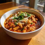 A close-up of a white bowl filled with Spicy Turkey White Bean Chili, topped with shredded cheese and fresh cilantro.