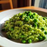 A close-up of creamy, bright green Spring Pea and Mint Risotto topped with whole peas and fresh herbs on a white plate.