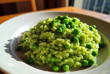A close-up of creamy, bright green Spring Pea and Mint Risotto topped with whole peas and fresh herbs on a white plate.