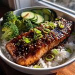 A close-up of a Teriyaki Salmon Rice Bowls serving featuring glazed salmon over white rice with broccoli and cucumbers.