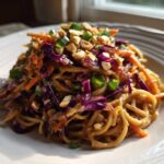 A serving of creamy Thai Peanut Noodles with Crunchy Veggies, topped with chopped peanuts and green onions on a white plate.