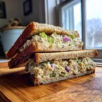 Two halves of a Vegan Chickpea Salad Sandwich stacked on a wooden cutting board, showing creamy filling with celery and red onion.